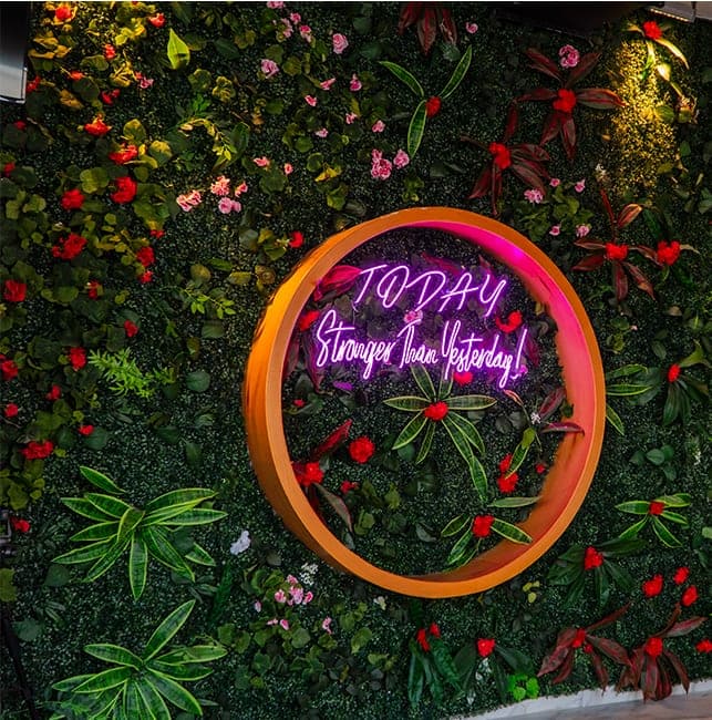 Yeşil bitki duvarı üzerinde turuncu çember içinde “Today — Stronger Than Yesterday!” neon yazısı