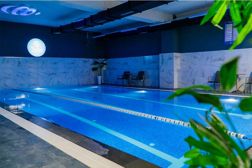 Today Living Club kapalı yüzme havuzu: mavi mozaik kaplı, kulvar ipiyle bölünmüş havuz ve duvarda ışıklı Today logosu