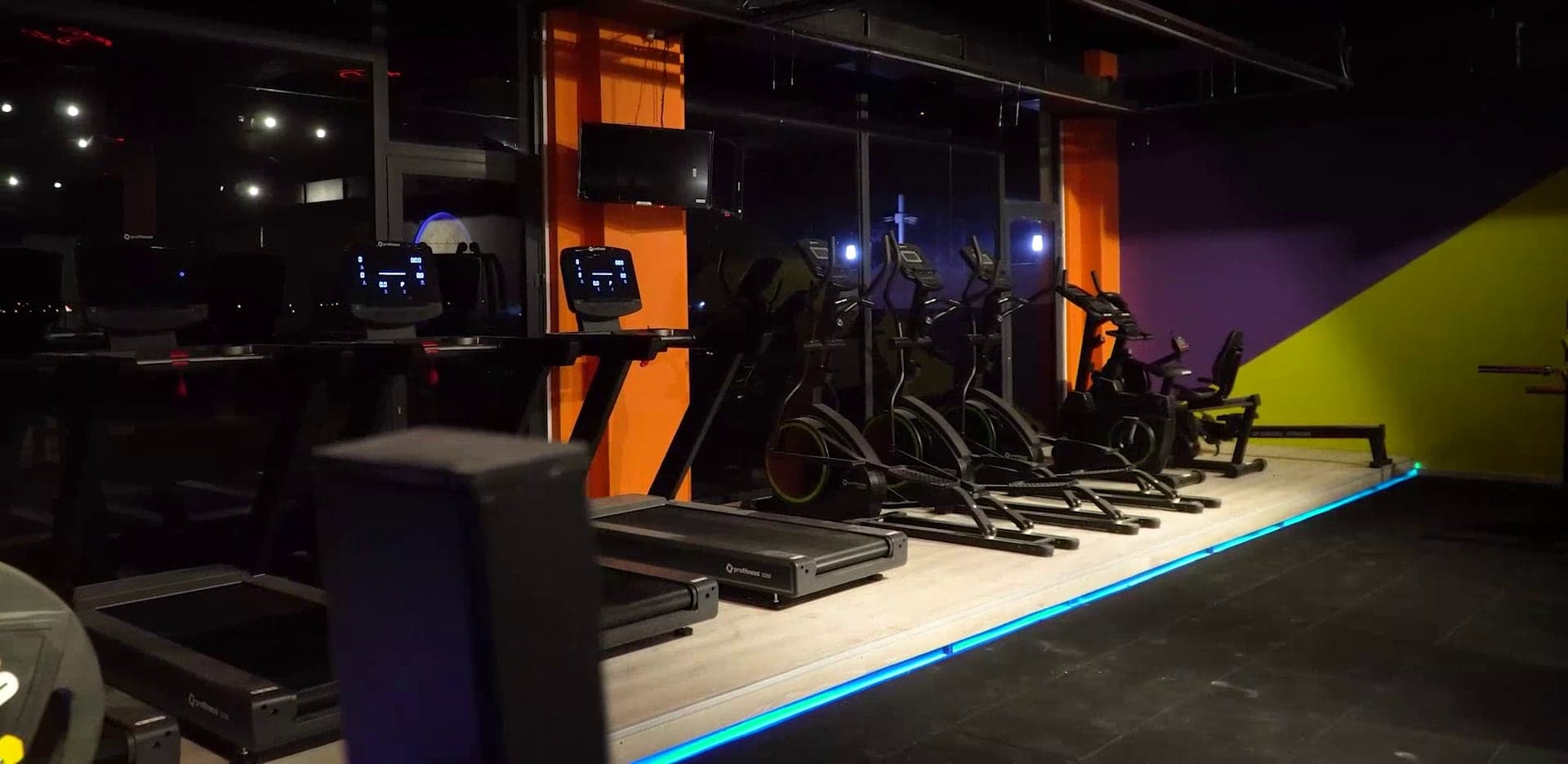 Mavi LED hattı boyunca dizili koşu bantları ve eliptikler, turuncu kolonlar
