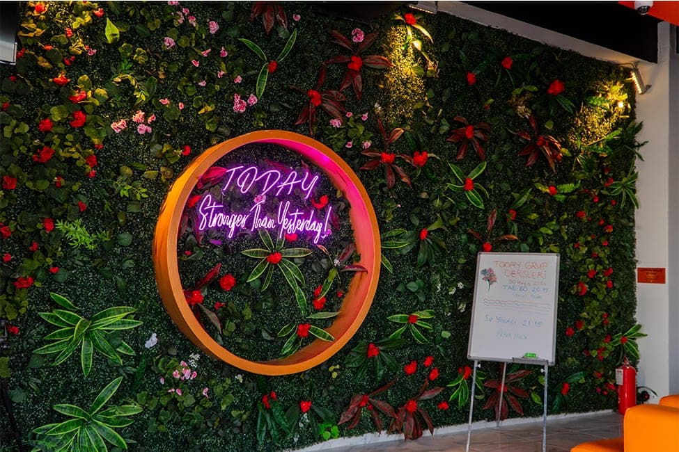 Yeşil bitki duvarı üzerinde turuncu çember içinde “Today — Stronger Than Yesterday!” neon yazısı