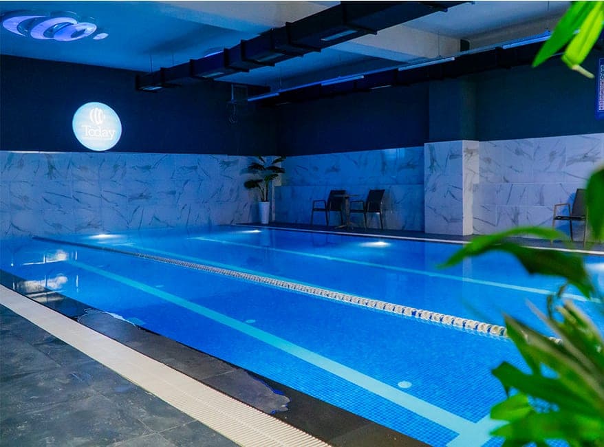Today Living Club kapalı yüzme havuzu: mavi mozaik kaplı, kulvar ipiyle bölünmüş havuz ve duvarda ışıklı Today logosu